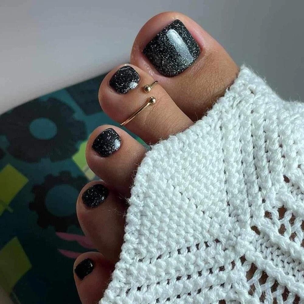 Czarny pedicure hybrydowy z brokatem i złotą obrączką na palcu